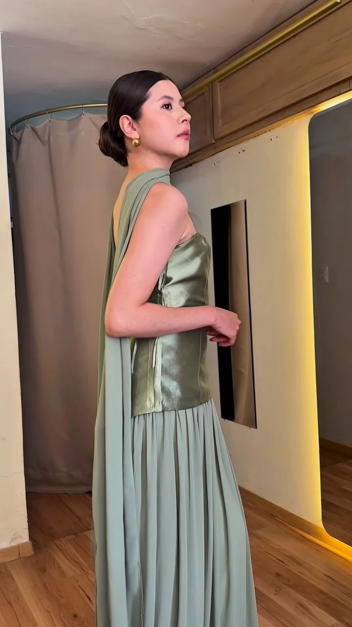 Una modelo con vestido largo verde salvia de satén, con capa al hombro y falda plisada, en el probador