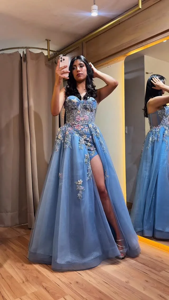 Una modelo frente al espejo con vestido largo azul de tul, con flores bordadas y abertura en la falda